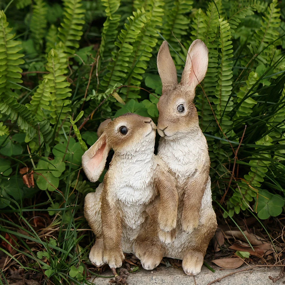 Two realistic brown and white resin rabbit figurines nestled among green garden ferns.
