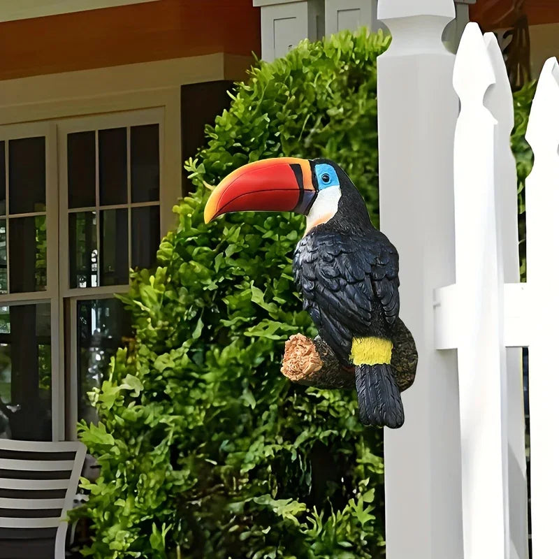 Decorative toucan figure on a branch in front of a white picket fence and green bush.