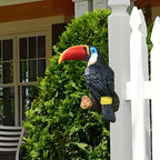 Decorative toucan figure on a branch in front of a white picket fence and green bush.