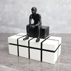 Matte black abstract long-legged thinker sculpture seated on a white textured cube box with black accent lines