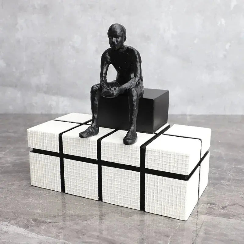 Matte black abstract long-legged thinker sculpture seated on a white textured cube box with black accent lines