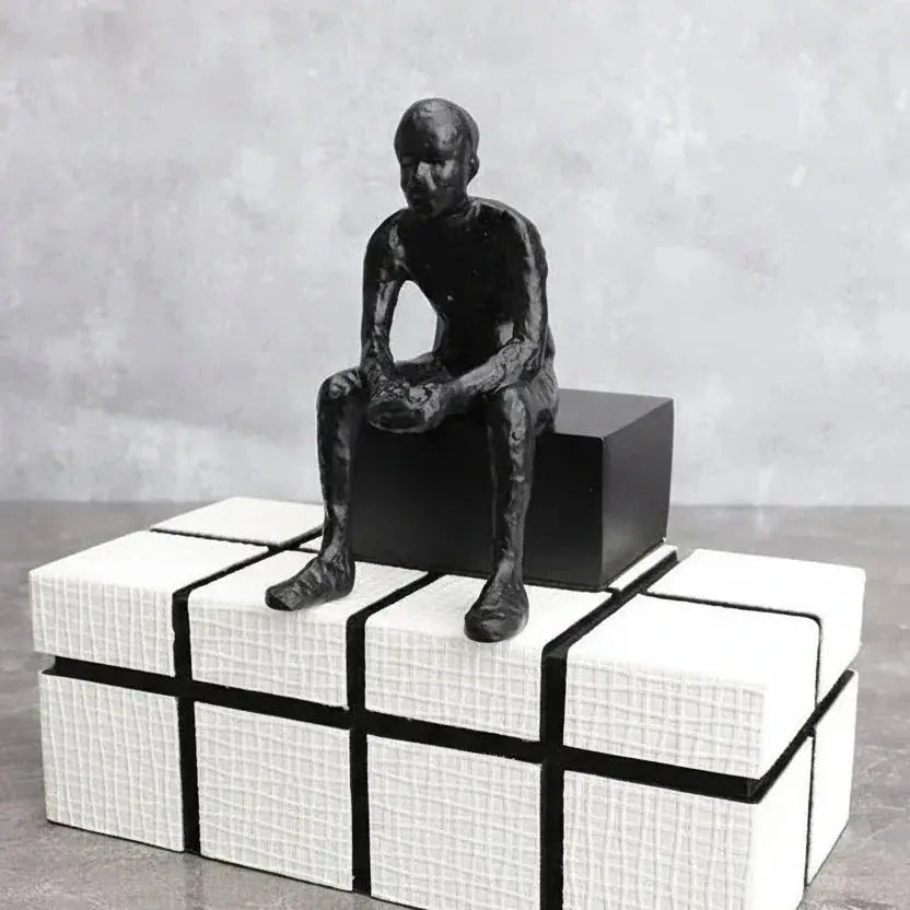 Modern matte black thinker sculpture of a seated man with clasped hands on white textured cubes with black lines